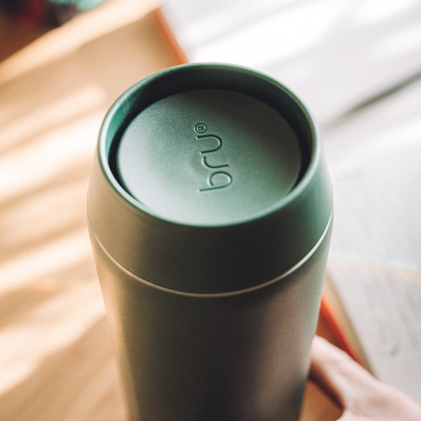 Overhead close-up of a green bru ceramic travel mug beside an open book in natural light
